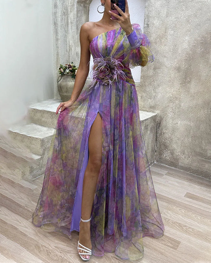 Kvinde i elegant asymmetrisk maxikjole med farverigt mønster, one-shoulder og slids. Ideel til sommerfest.